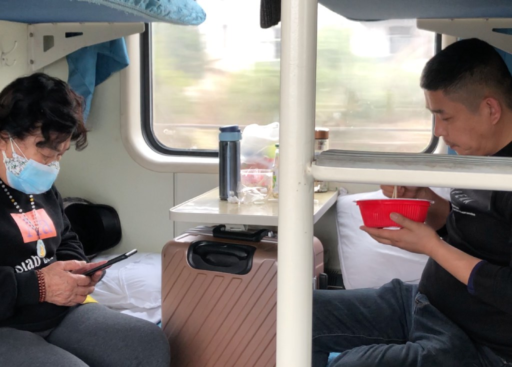 เตียงชั้นล่างของรถไฟจีน ทางซ้ายมีผู้หญิงนั่งเล่นโทรศัพท์มือถือ ทางขวามีผู้ชายนั่งกินอาหาร มีโต๊ะเล็กอยู่ตรงกลาง